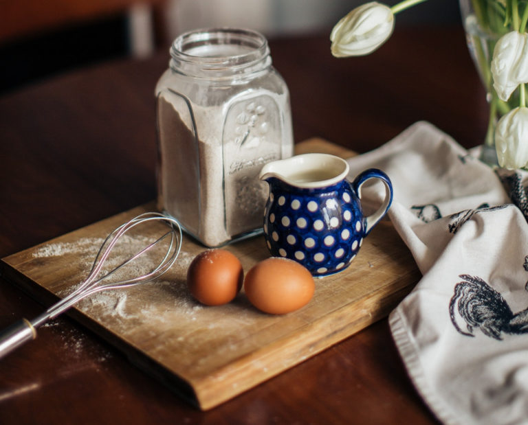 two brown eggs, whisk, blue & white polka dot pitcher and glass jar containing flour sitting on wooden cutting board